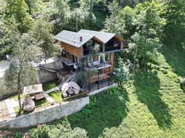 Una casa moderna de varios pisos con un muro de contención de piedra, terraza de madera y un ventanal con vistas a un jardín exuberante.