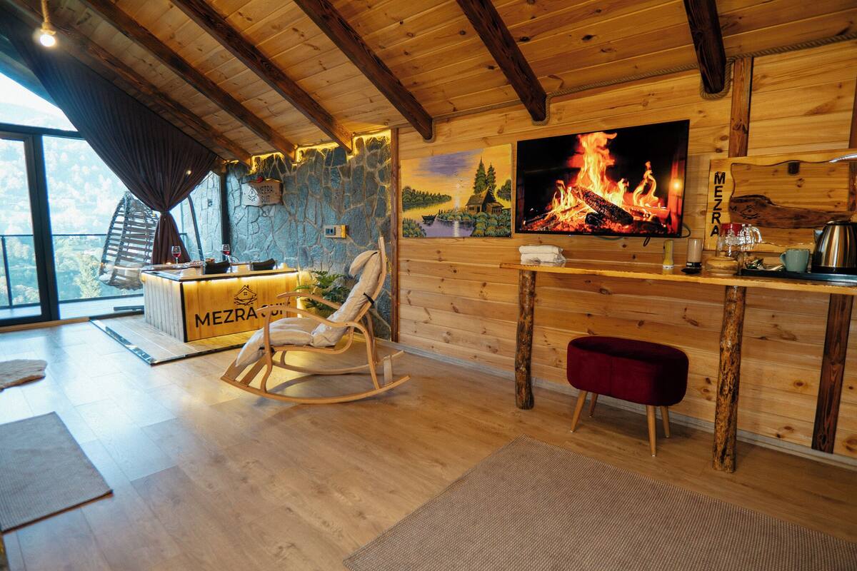 A cozy room with a stone wall, wooden ceiling, a rocking chair, a red bench, a TV displaying a fire, and a bar area with a 'MEZRA' sign.