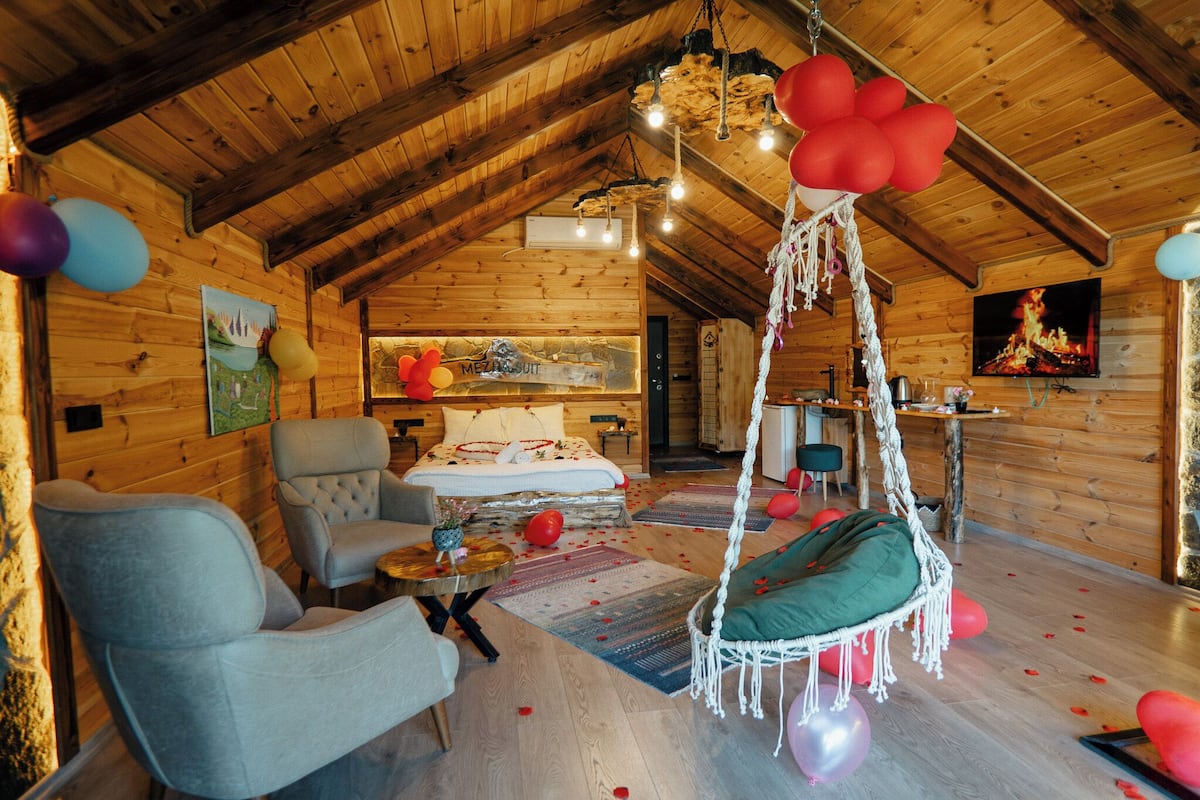 A cozy log cabin interior with a wooden ceiling, a bed with a white comforter, a hanging chair, and a dining area with a table and chairs.