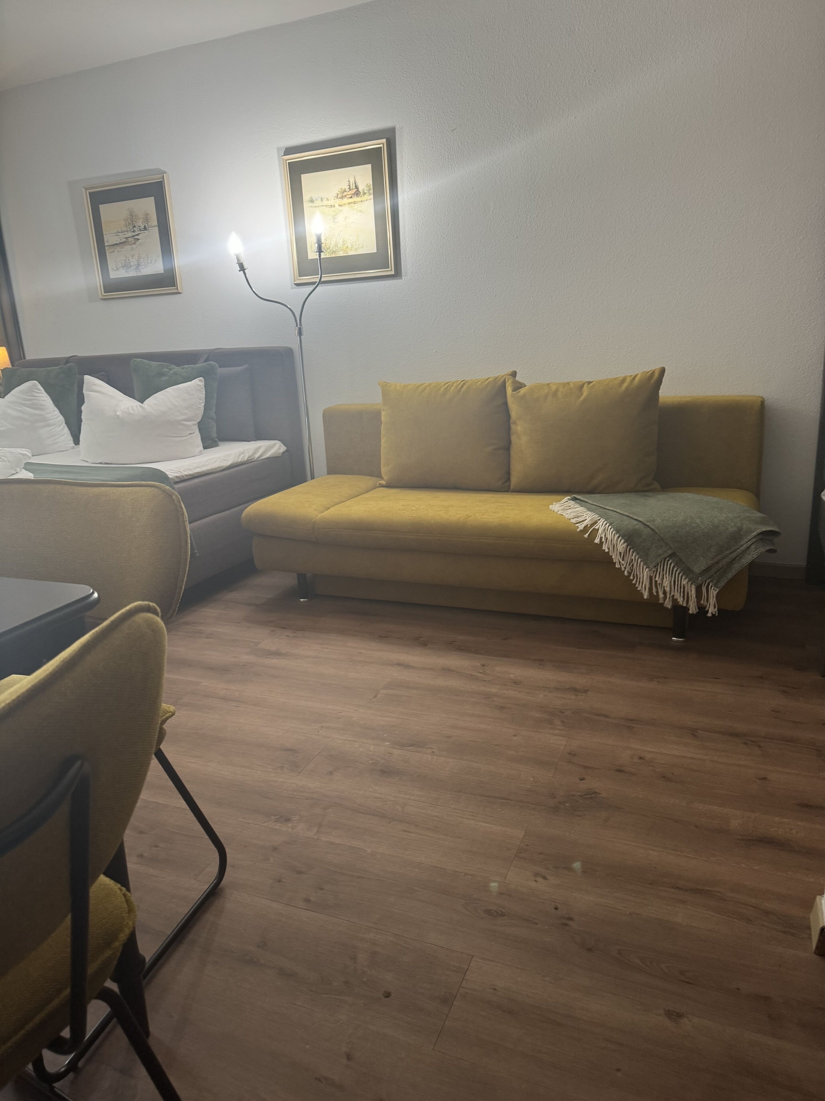 A room with a yellow sofa, a small table, and a lamp. There are two framed pictures on the wall.