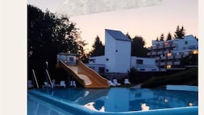 A swimming pool with a slide, surrounded by buildings and trees. - Stressless Hotelapparat Inklusive Aktivcard Bayerischer Wald (Sankt Englmar)
