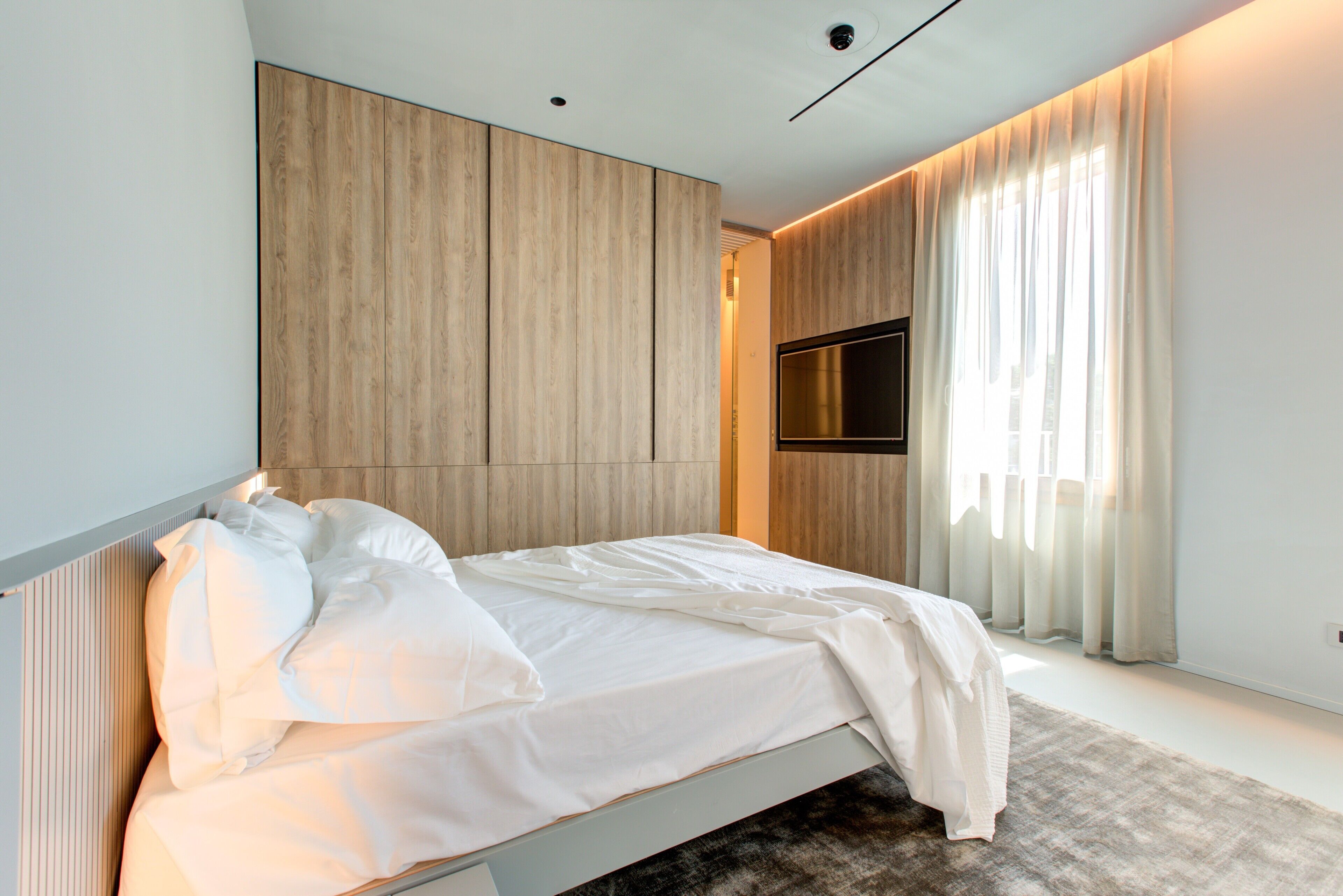A modern bedroom with a bed, wooden panel wall, TV, and large window with curtains.