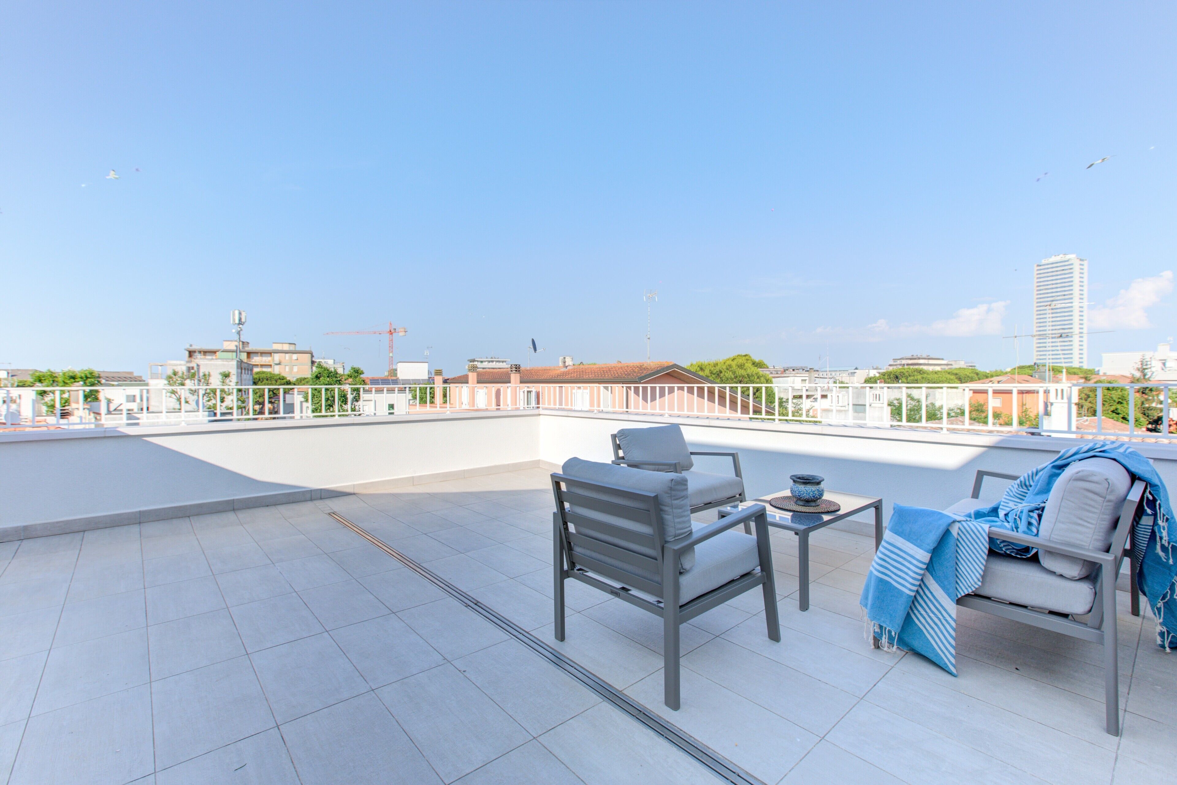 A rooftop terrace with a seating area, including a table and chairs, overlooking a cityscape with buildings and a clear sky.