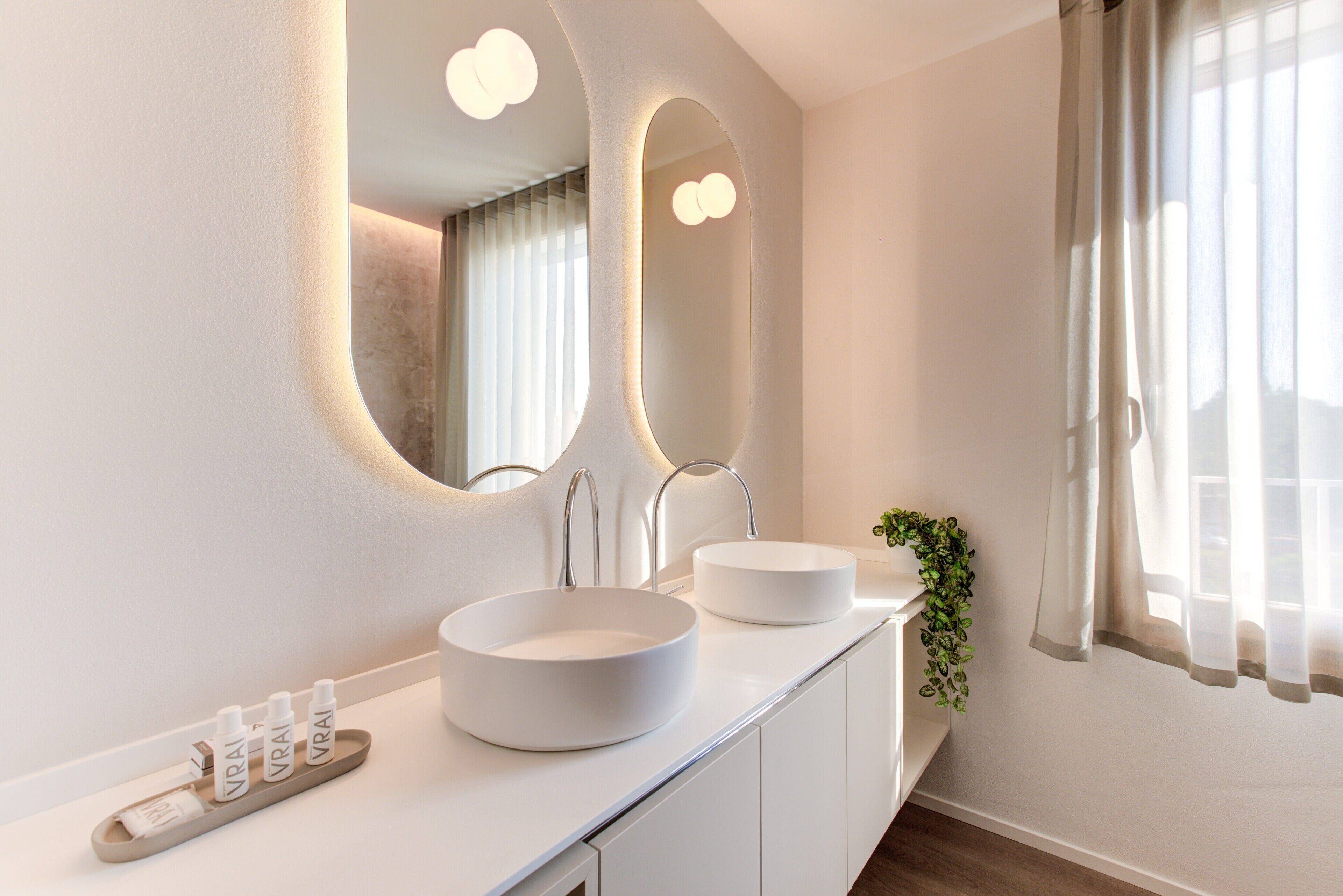 Un baño moderno con dos lavamanos redondos blancos, un espejo grande con iluminación integrada y una planta en maceta sobre la encimera.
