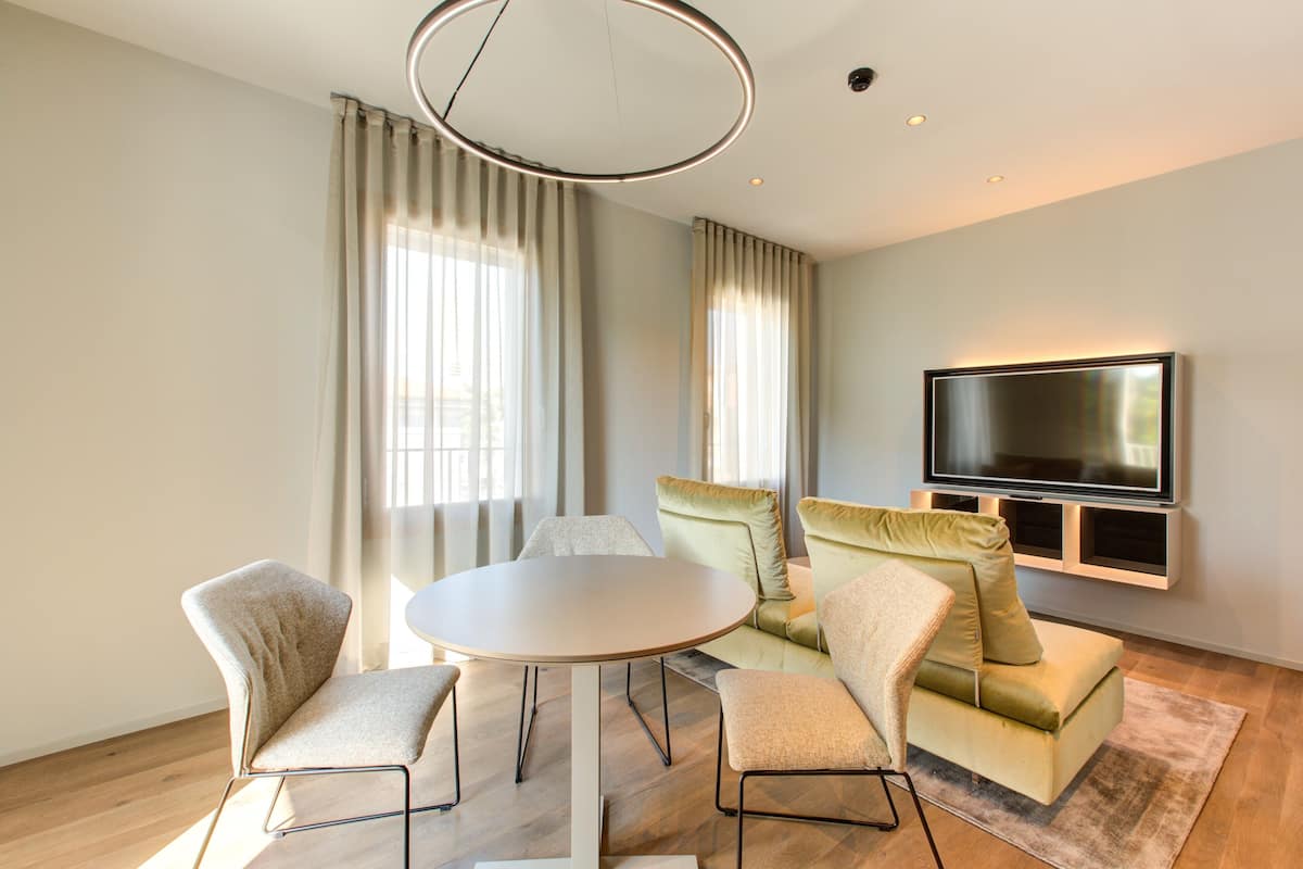 Una sala de estar moderna con una mesa redonda, dos sillones, un sofá, un televisor de pantalla plana montado en la pared y ventanas grandes con cortinas.