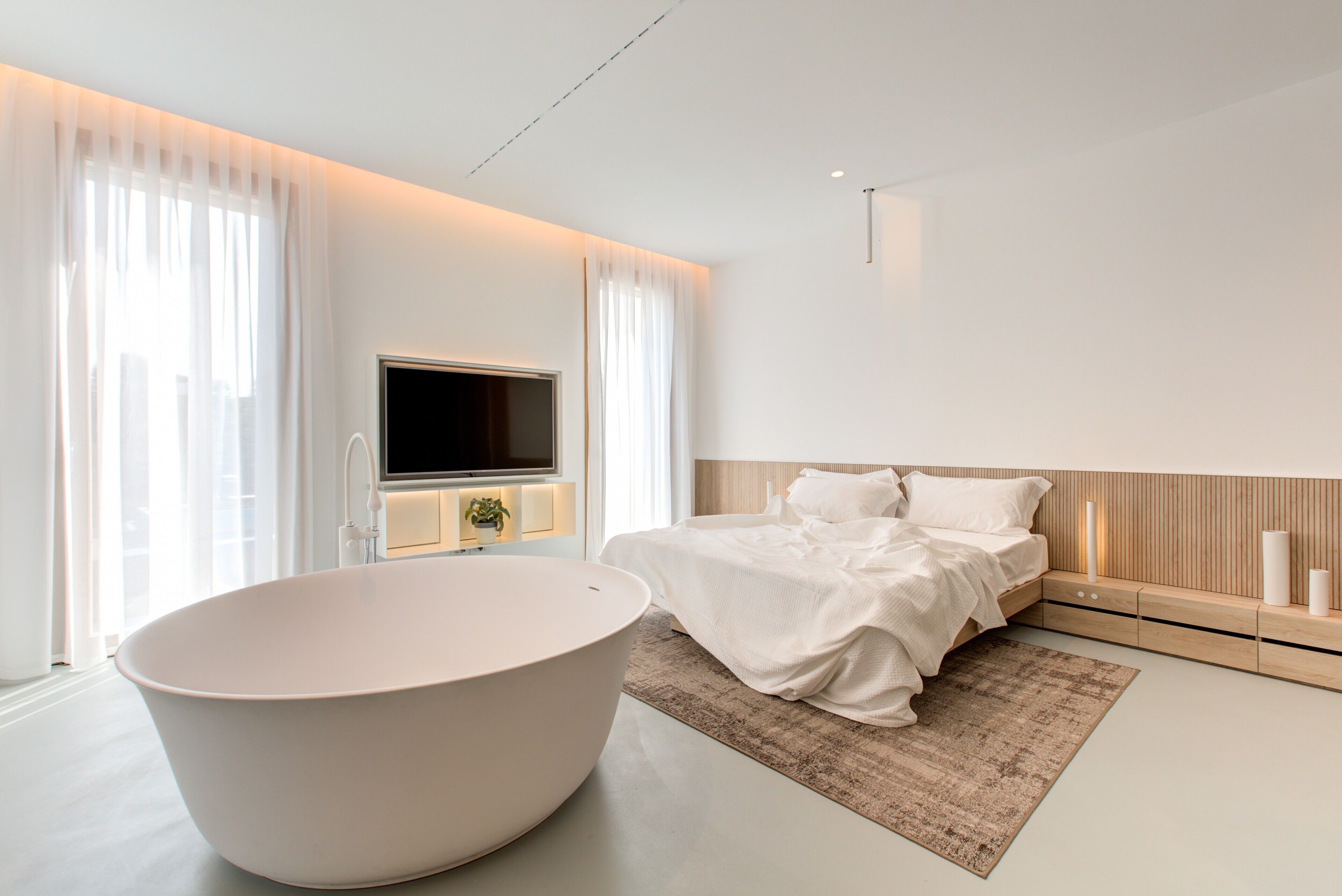 Een moderne slaapkamer met een ruime badkuip, een bed met witte lakens, een flatscreen televisie aan de wand en een nachtkastje met een lamp.