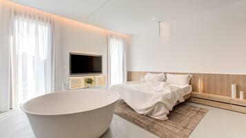 Een moderne slaapkamer met een ruime badkuip, een bed met witte lakens, een flatscreen televisie aan de wand en een nachtkastje met een lamp.