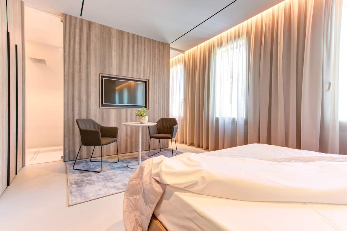 Una habitación de hotel moderna con una cama grande, un televisor de pantalla plana montado en la pared, dos sillas alrededor de una mesita pequeña y un ventanal con cortinas.