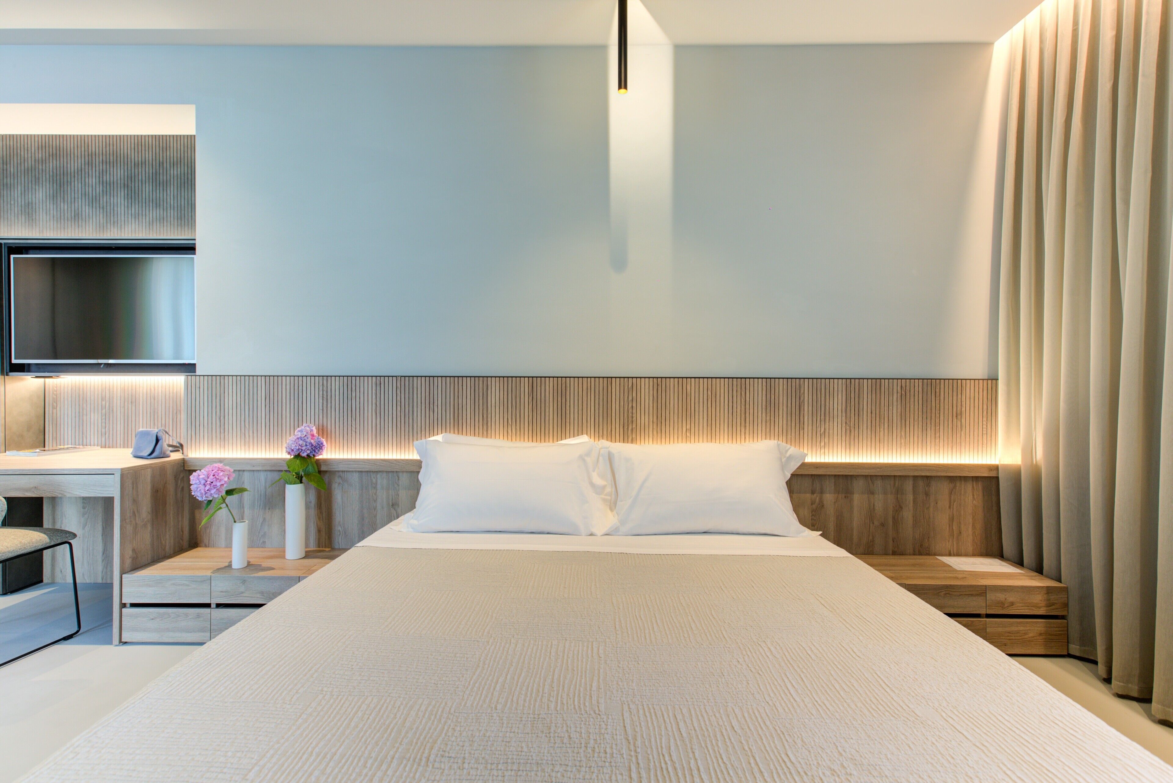 A modern hotel room with a large bed, a desk with a chair, a flat-screen TV, and a bedside table with two vases of flowers.