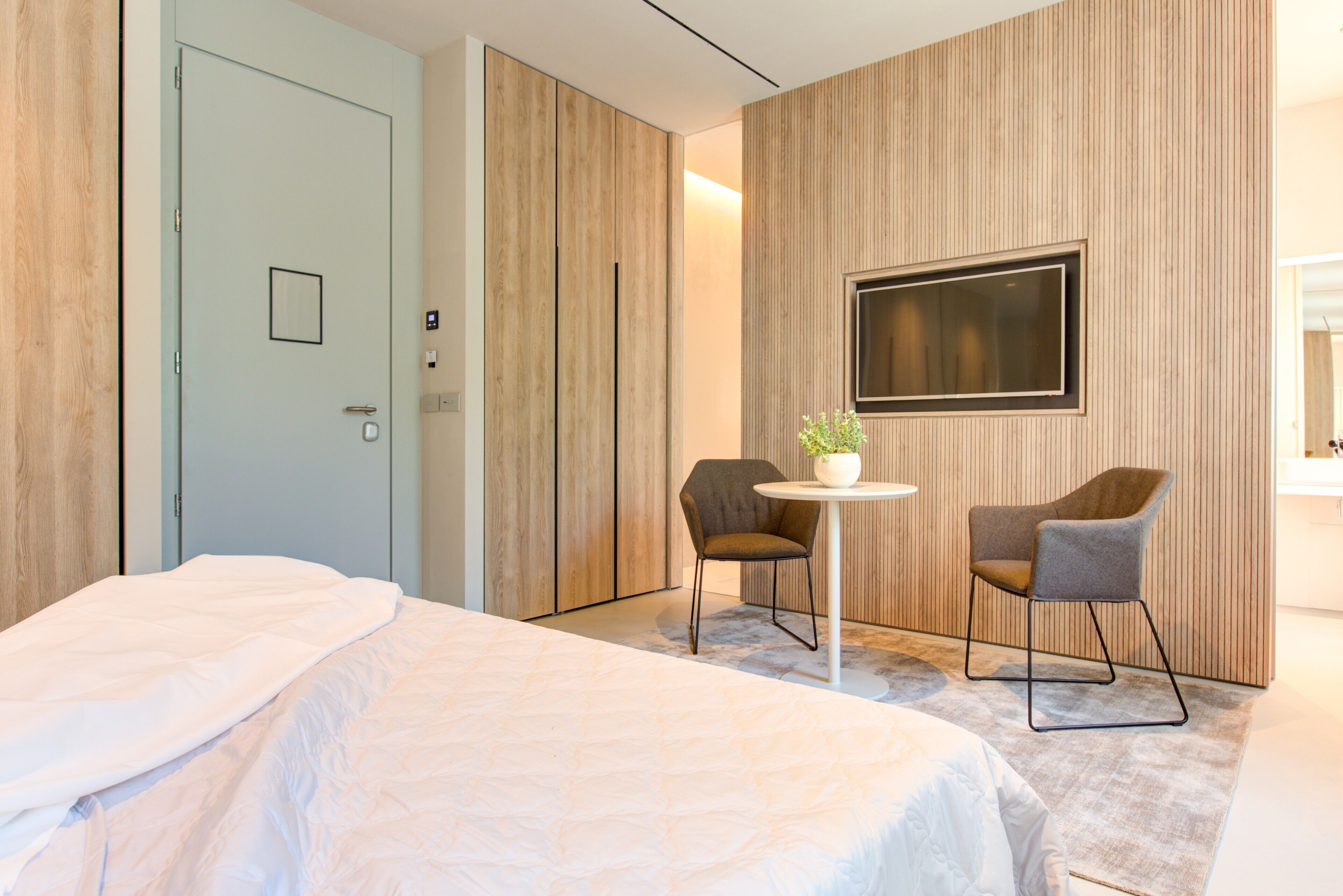A modern hotel room with a large bed, a small round table with two chairs, a potted plant, and a TV mounted on the wall.