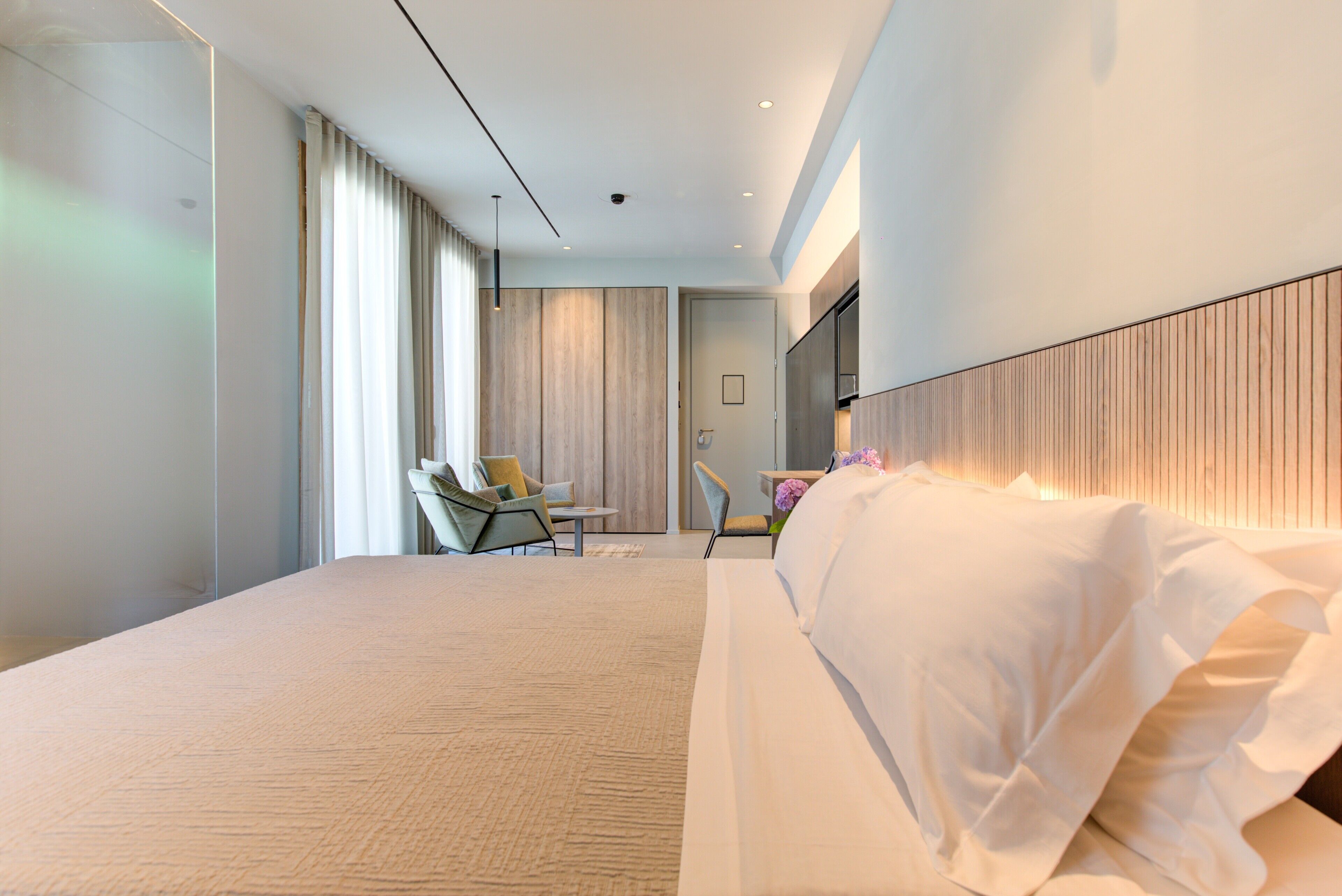 Una habitación de hotel moderna con una cama grande, un área de descanso con una silla y una mesa, y un ventanal con cortinas.