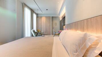 Una habitación de hotel moderna con una cama grande, un área de descanso con una silla y una mesa, y un ventanal con cortinas.
