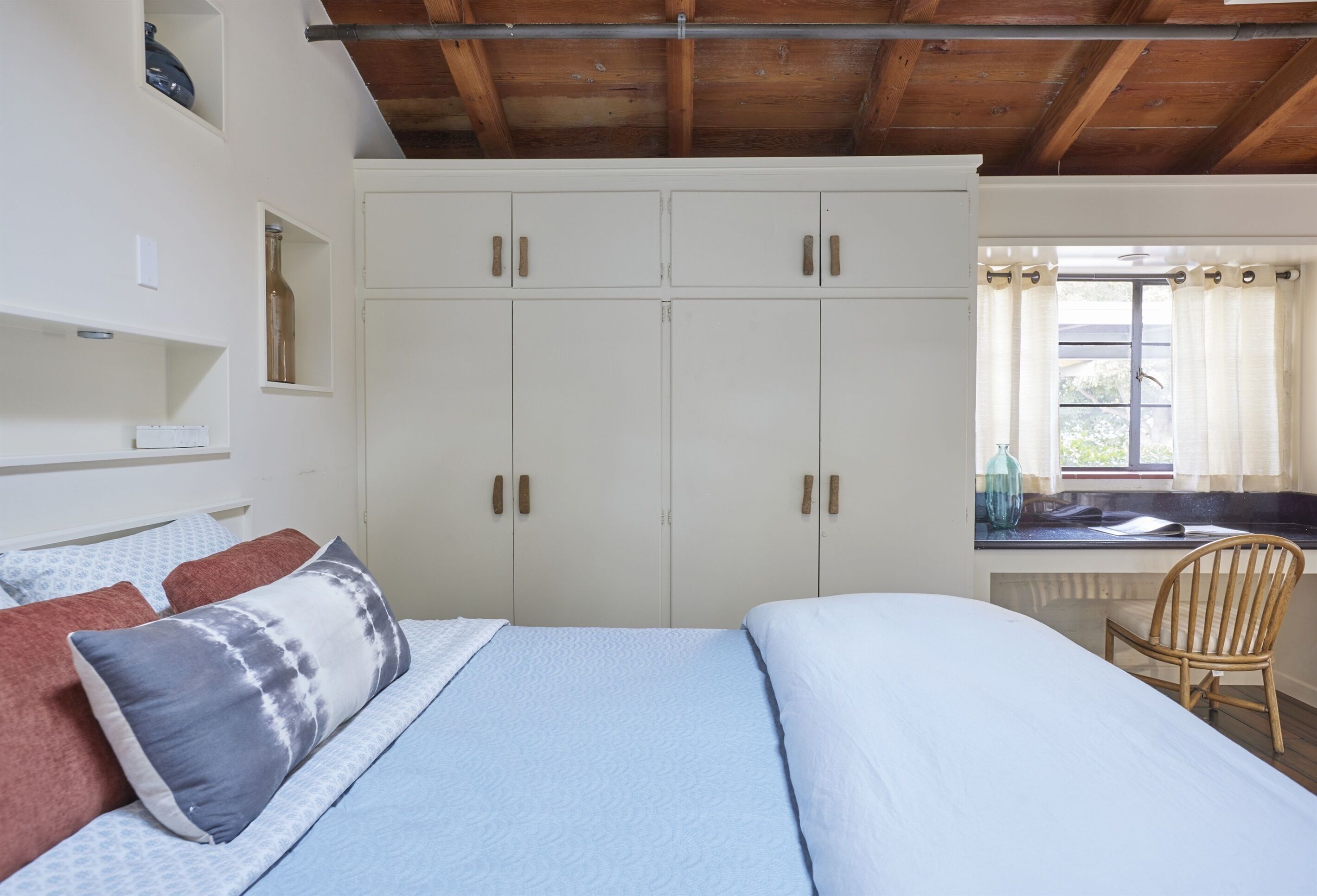 Un dormitorio con cama, almohadas, una silla de madera, una ventana con cortinas y un armario blanco.