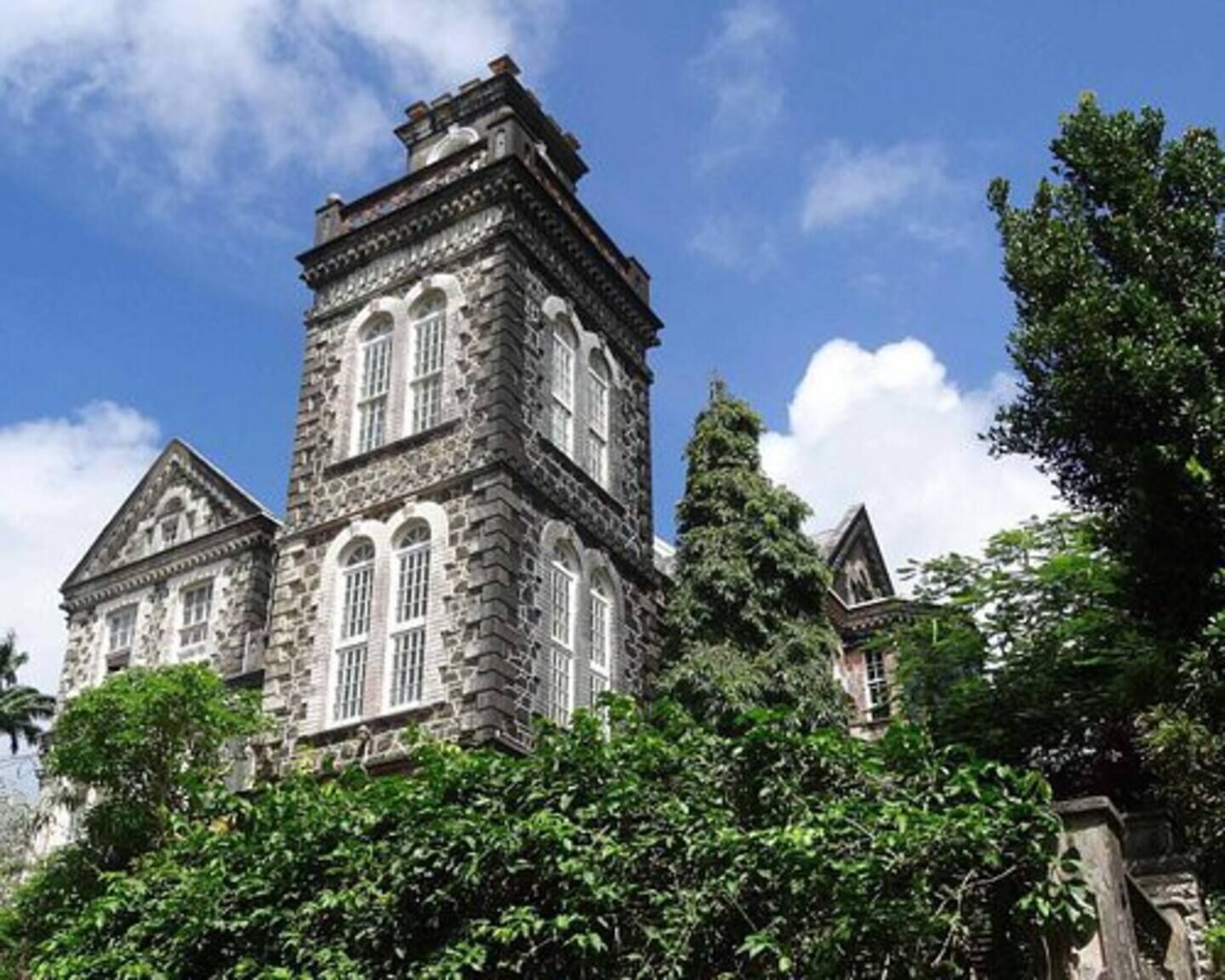 A historic stone building with a prominent tower and arched windows, surrounded by lush greenery.