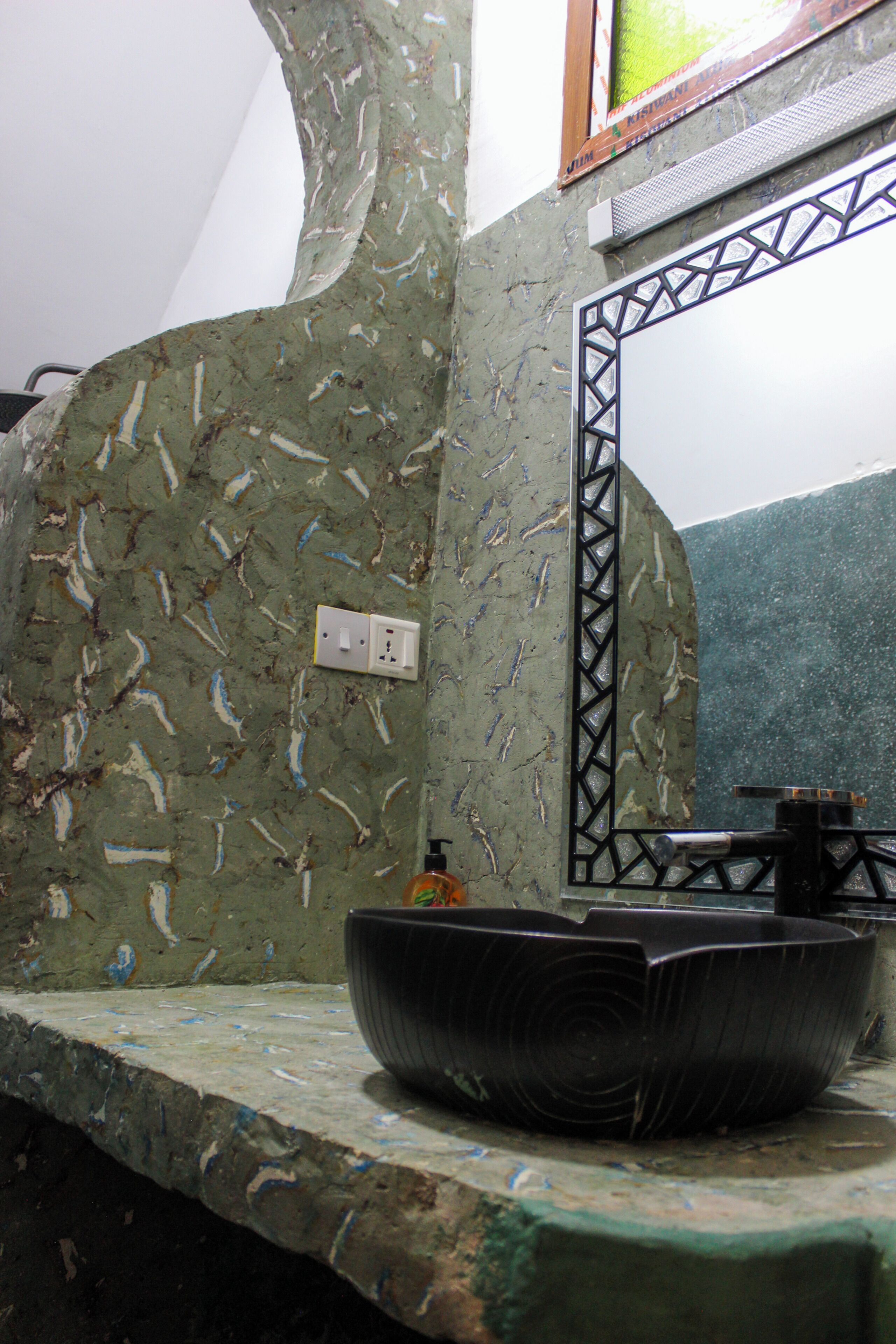 A bathroom with a unique textured wall, a sink with a black basin, and a mirror with a geometric frame.