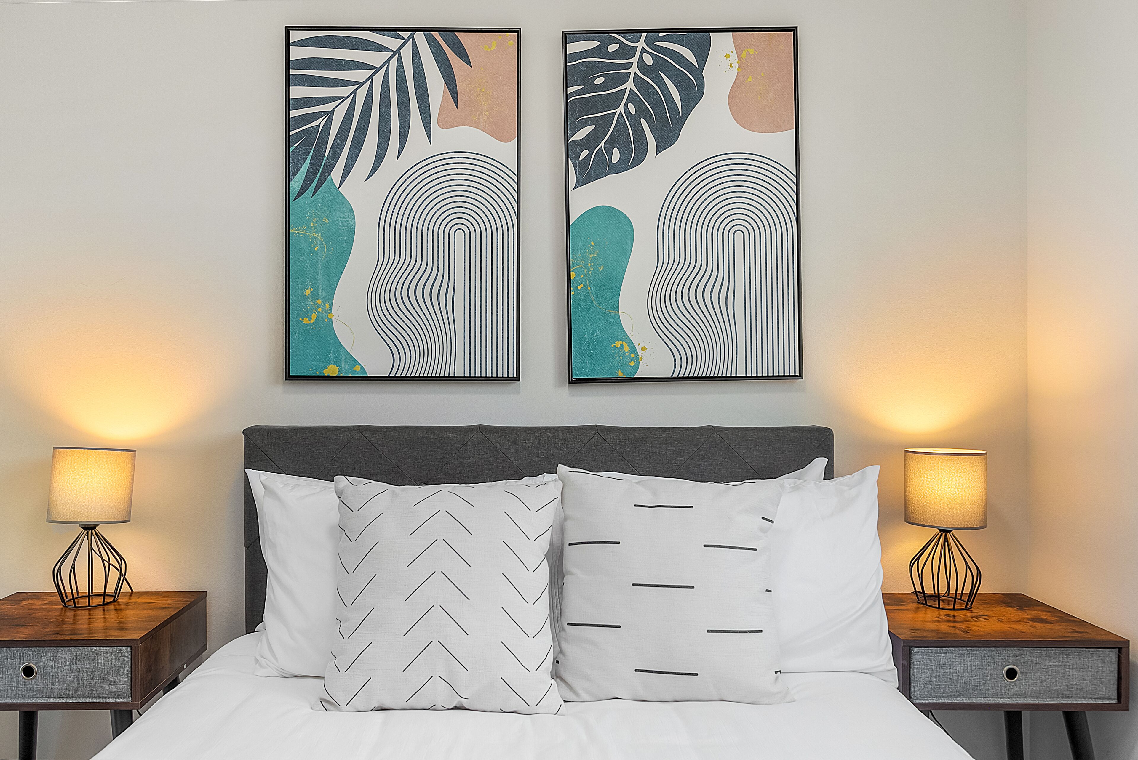 Un lit bien fait, avec deux oreillers, flanqué de deux tables de chevet sur lesquelles reposent des lampes, et deux œuvres d’art accrochées au mur au-dessus.