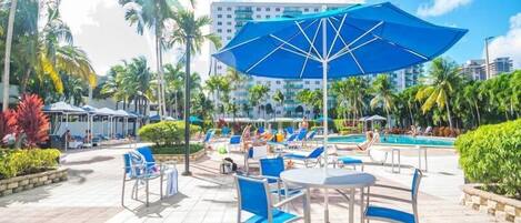 A poolside area with lounge chairs and umbrellas, surrounded by palm trees and a building in the background.