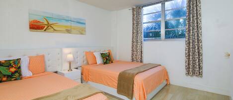 Un dormitorio con dos camas, una ventana con cortinas, una mesita de noche con lámpara y un cuadro en la pared.