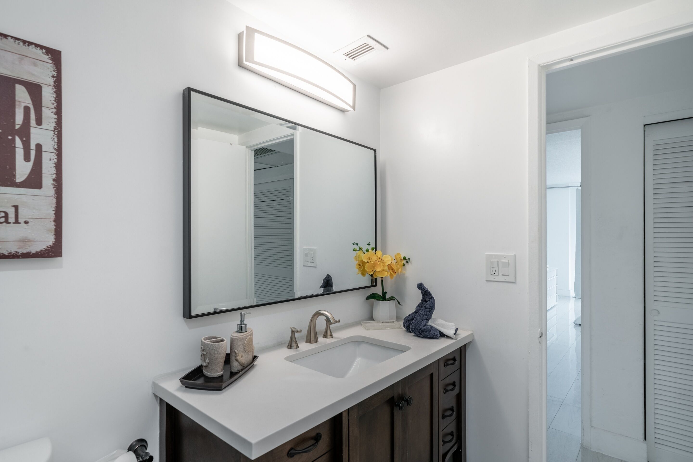 A modern bathroom with a white sink, a large mirror, and a small vase with yellow flowers.