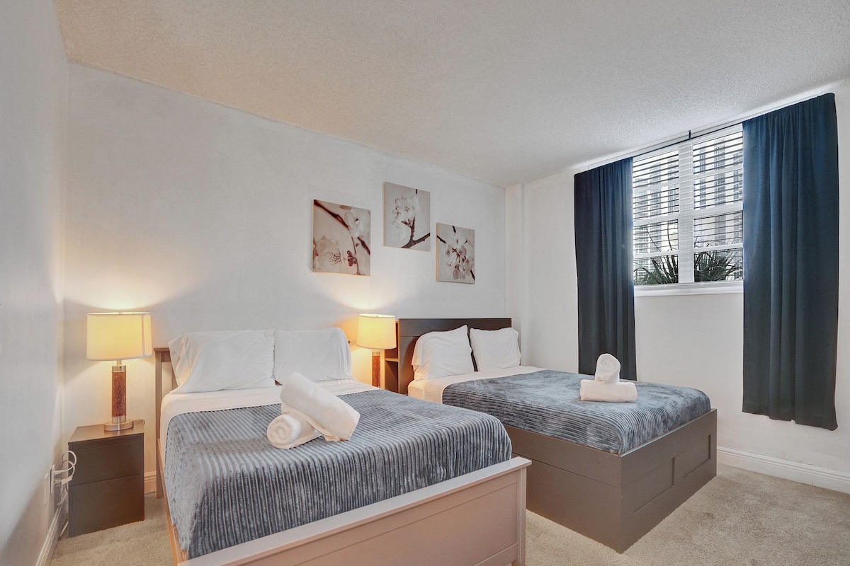 A hotel room with two beds, a window with curtains, and artwork on the wall.