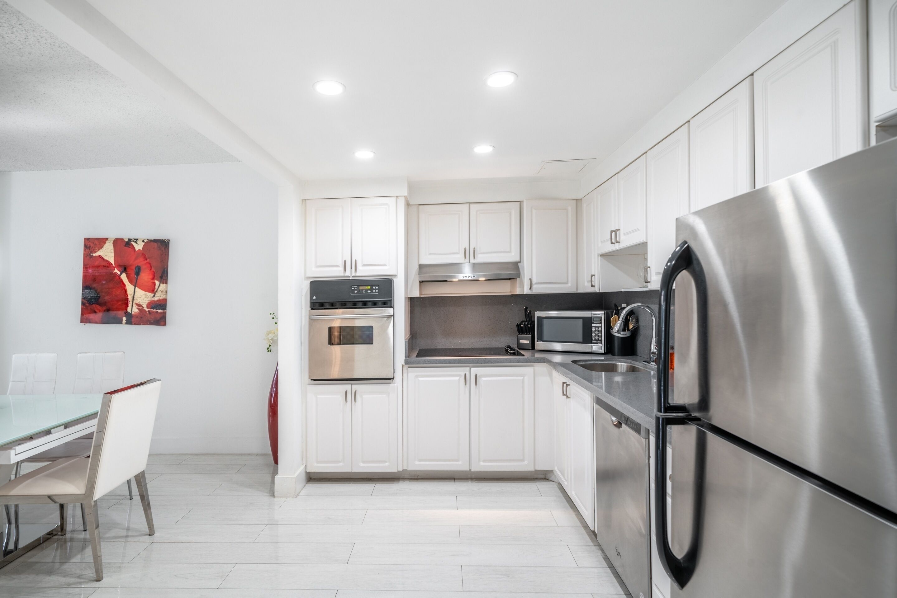 Una cocina moderna con gabinetes blancos, electrodomésticos de acero inoxidable y un comedor con mesa y sillas de vidrio.