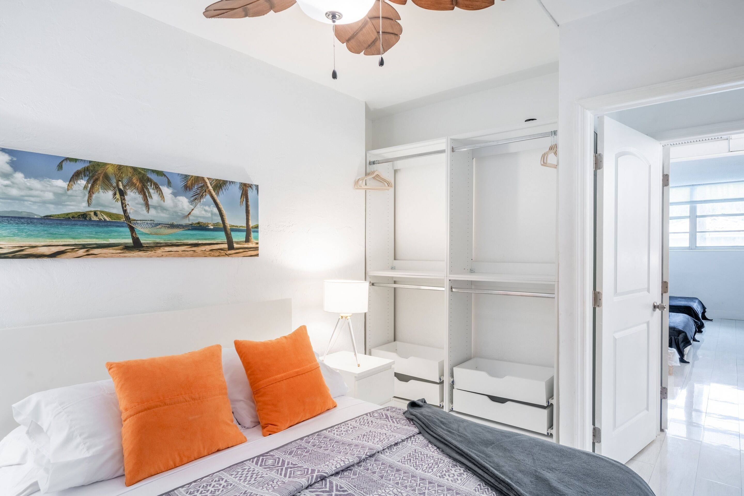 Een slaapkamer met een bed, oranje kussens, een nachtkastje, een lamp, een kledingkast en een schilderij van een strandgezicht.