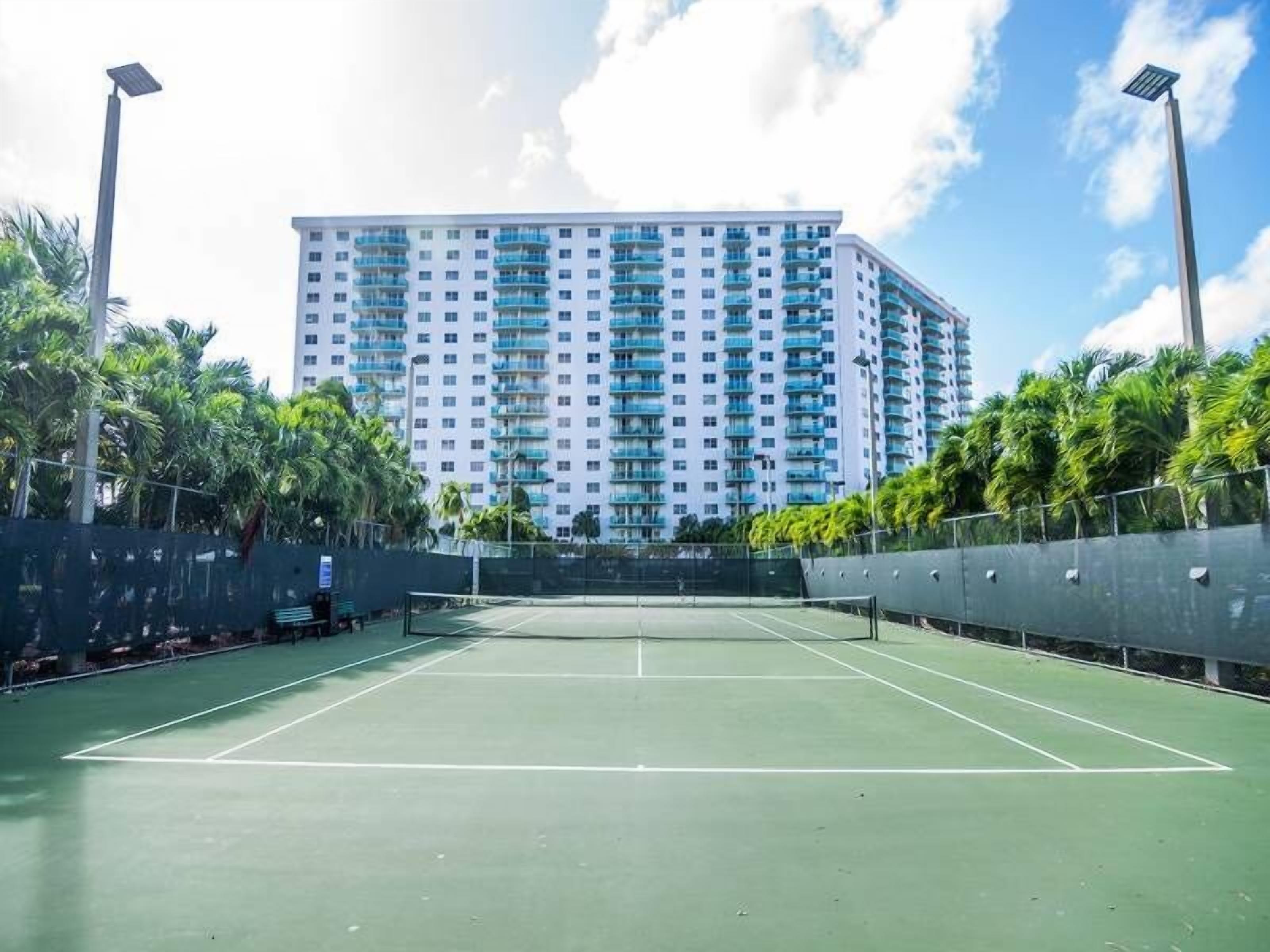 Un court de tennis en plein air, doté d’un filet et entouré d’une clôture, avec un grand immeuble en arrière-plan.