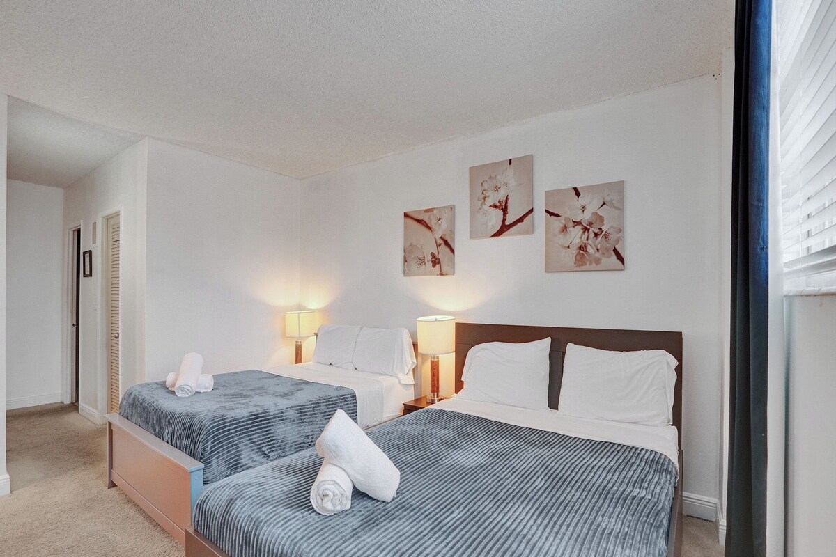 A hotel room with two beds, a door, a lamp, and wall art.