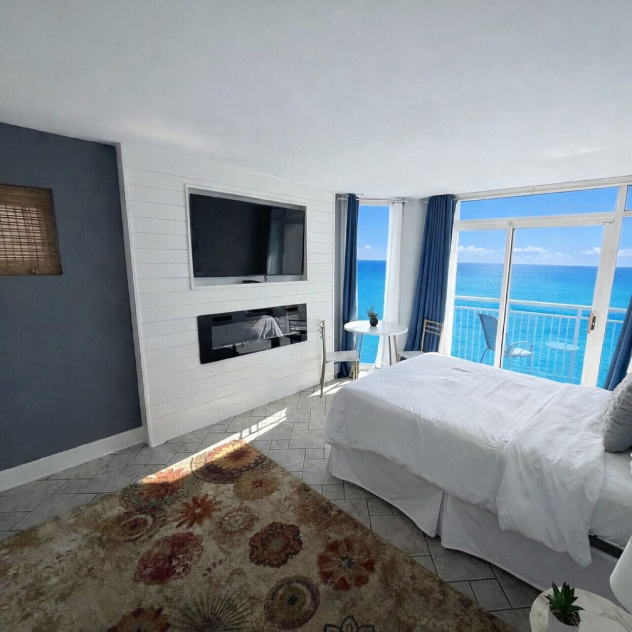 Quarto com cama, televisão na parede, mesinha com cadeiras e varanda com vista para o mar.