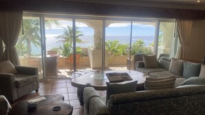 Un vaste espace de vie avec une grande porte-fenêtre coulissante donnant sur un balcon avec vue sur l’océan. La pièce est aménagée avec plusieurs canapés et une table basse.