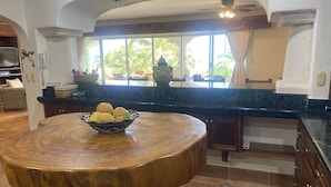 Une cuisine avec une table ronde en bois, un saladier rempli de fruits et, par la fenêtre, une vue sur des palmiers.