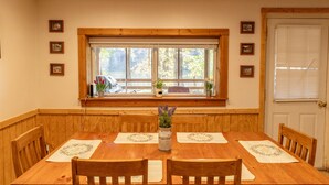 A dining room with a wooden table, four chairs, a window with a view of trees, and a door with blinds. - Escape to the Shenandoah River and Relax (Luray)