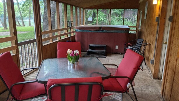 A covered patio with a glass table and red chairs, a hot tub, and a view of a lake. - Escape to the Shenandoah River and Relax (Luray)