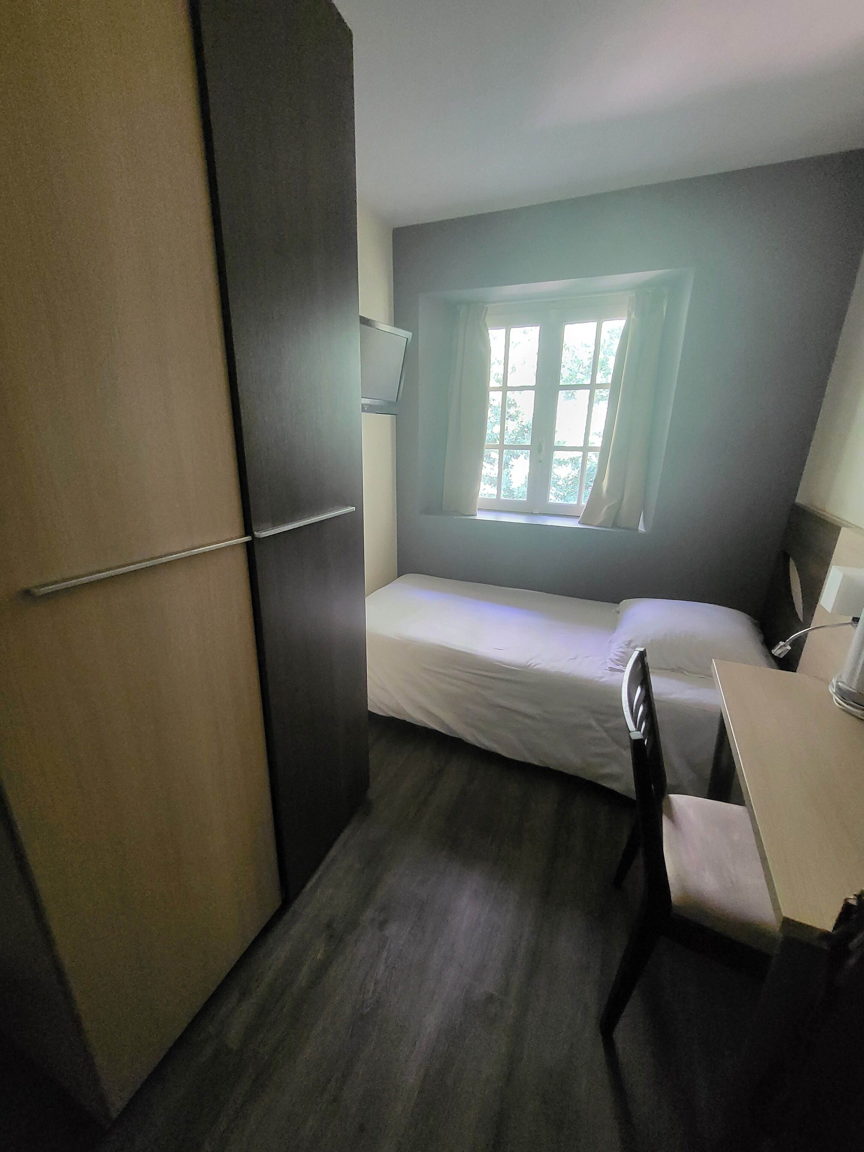 A small, compact room with a single bed, a desk, and a window with curtains.