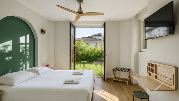 Un dormitorio con cama, ventilador de techo, televisor montado en la pared y vista a un jardín a través de una puerta abierta.