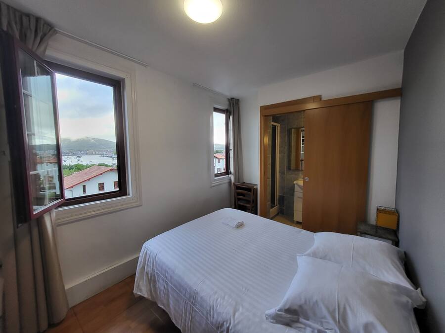 Un dormitorio con cama, ventana con vistas a edificios y montañas, armario de madera y baño con espejo y ducha.