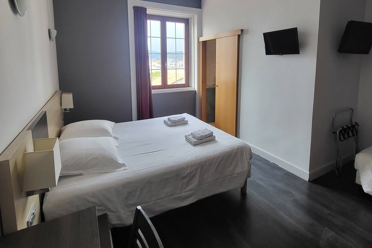 A hotel room with a bed, bedside tables, a desk, a chair, a window with curtains, a door, and a flat-screen TV mounted on the wall.