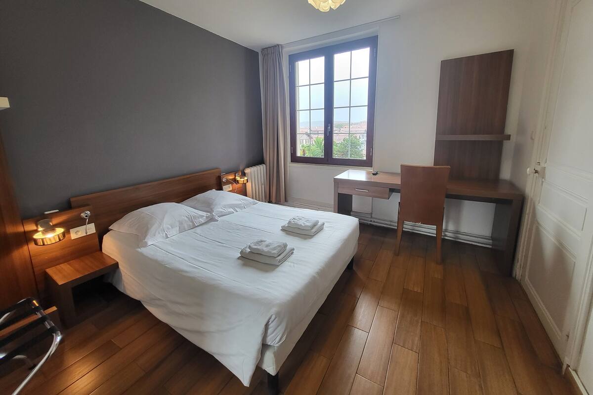 A hotel room with a bed, bedside table, lamp, desk, chair, and a view through the window.