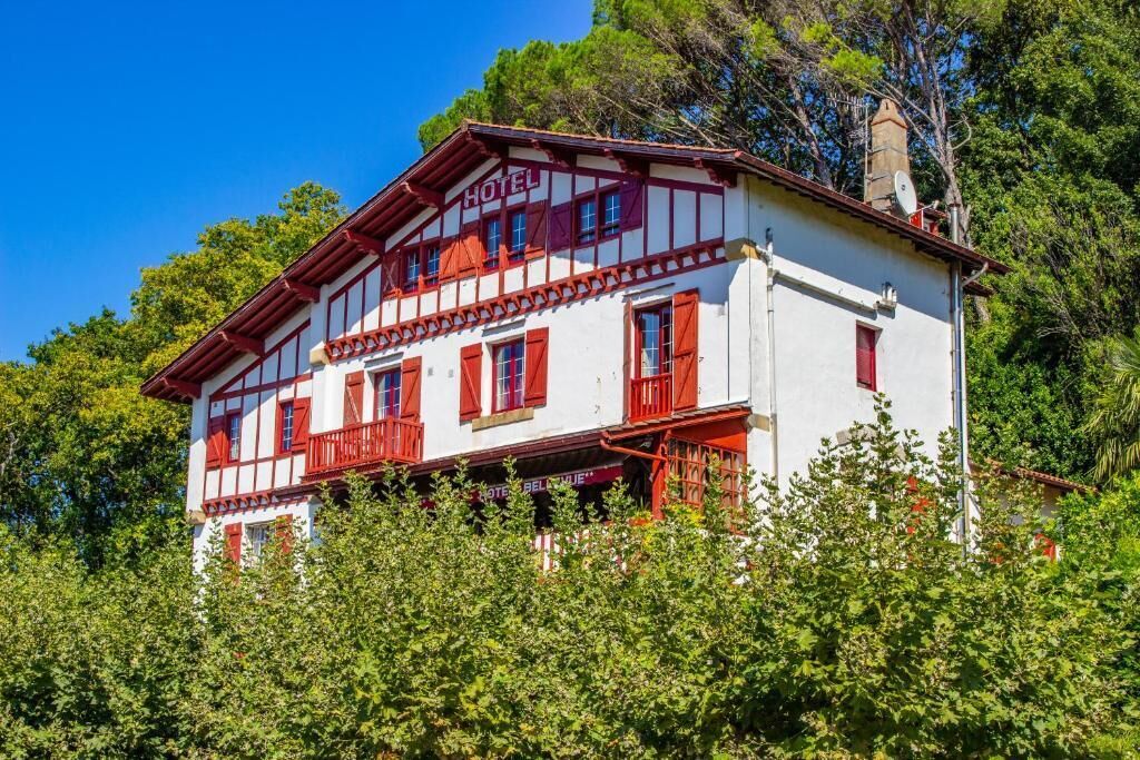 A two-story hotel with a white facade and red accents, surrounded by lush greenery.