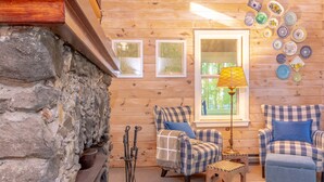 Interior acogedor de una cabaña con chimenea de piedra, sillones a cuadros, una mesita y una lámpara.