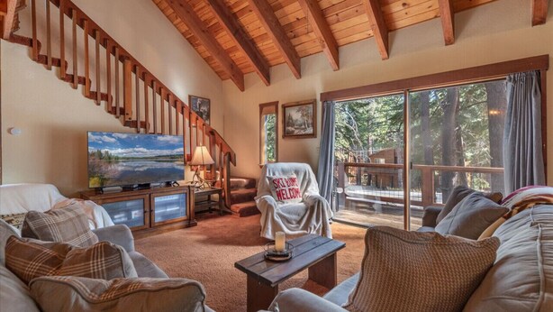 A cozy living room with a wooden staircase, a television, and a large window with a view of trees.