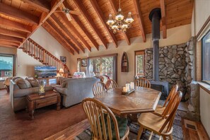 A spacious living area with a wooden ceiling, a stone fireplace, a dining table with chairs, and a staircase leading to another level.