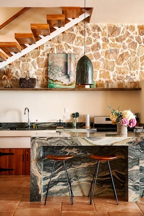 A kitchen with a stone wall, wooden staircase, marble island, and bar stools.