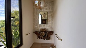 Ein Badezimmer mit Steinwand, Waschbecken aus Holz, einem Spiegel und einem großen Fenster mit Blick ins Grüne.
