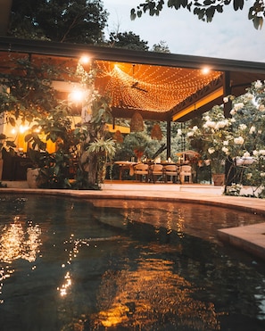 Área de piscina externa com luzes de corda, assentos e plantas em vasos.