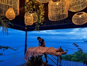 En massagebehandling foregår på en træterrasse med udsigt over havet.