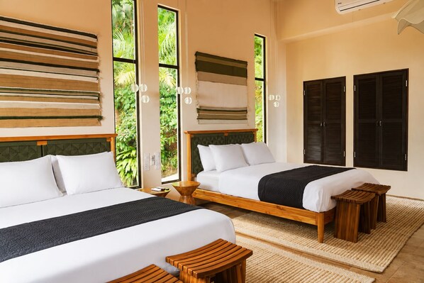 A hotel room with two beds, a large window with a view of greenery, and a wall-mounted air conditioning unit.