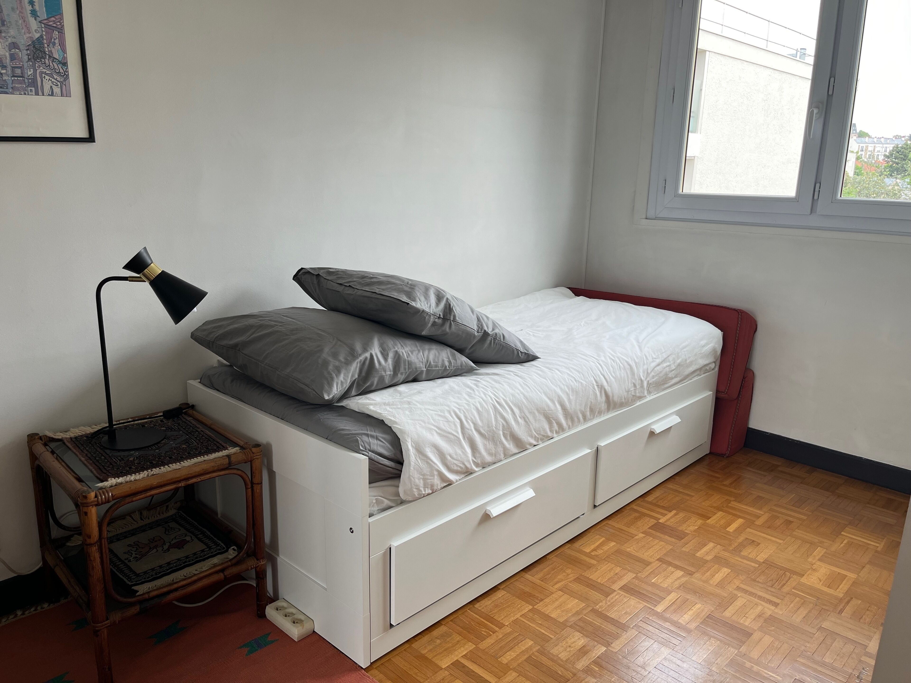 Habitación individual con una cama y cabecera blanca, ropa de cama gris, una mesita de noche con lámpara y ventana con vista a edificios.
