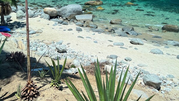 A beach with clear turquoise water, pebbles, and sparse vegetation. - Beach Front Condo with Outstanding Views  (Los Barriles)