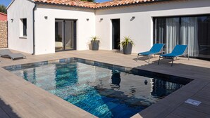 En moderne udendørs swimmingpool med klart, blåt vand, omgivet af en træterrasse og to blå liggestole.
